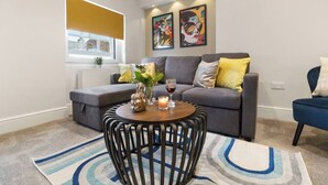 Una sala de estar moderna con un sofá gris, una mesa de centro redonda y un sillón azul. Hay dos cuadros enmarcados en la pared y una ventana con una persiana amarilla.