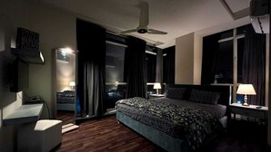 Une chambre d’hôtel avec un grand lit, un bureau, une chaise, un ventilateur de plafond et une fenêtre avec des rideaux.