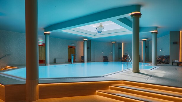 A modern indoor swimming pool with a skylight, large columns, and a central staircase.