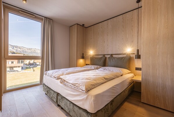 A modern bedroom with a large bed, wooden panel walls, and a view of a beach and buildings. - Nice Apartment With Shared Sauna (Oberndorf in Tirol)