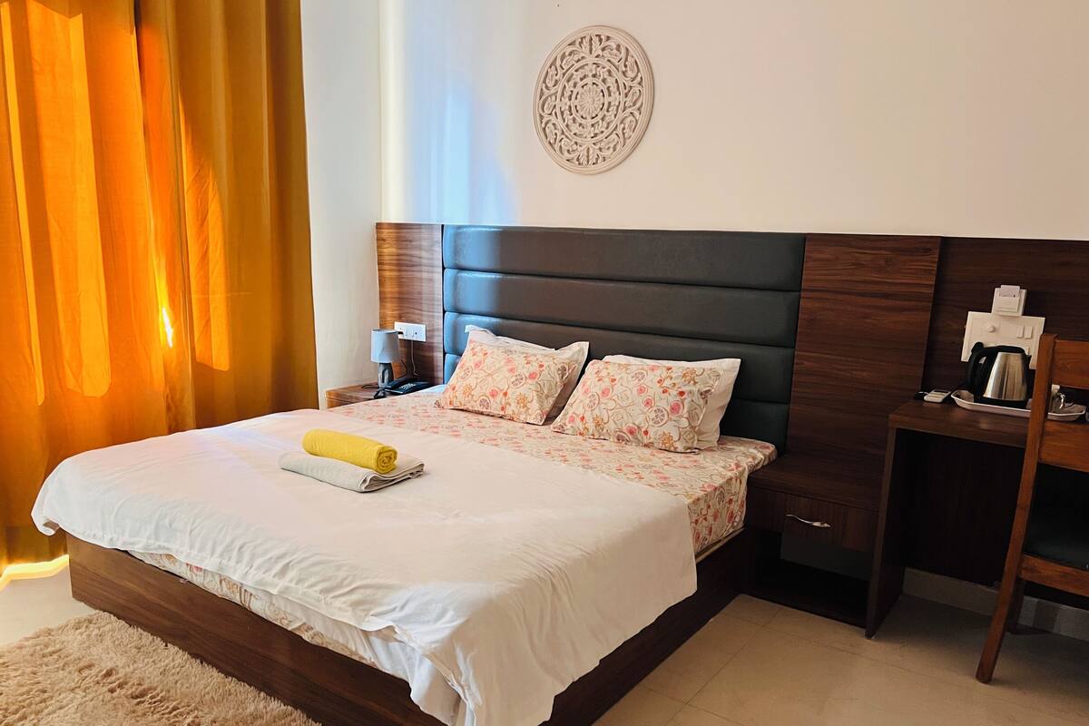 Cama bem arrumada com uma toalha enrolada, mesa de cabeceira com uma chaleira e uma luminária.