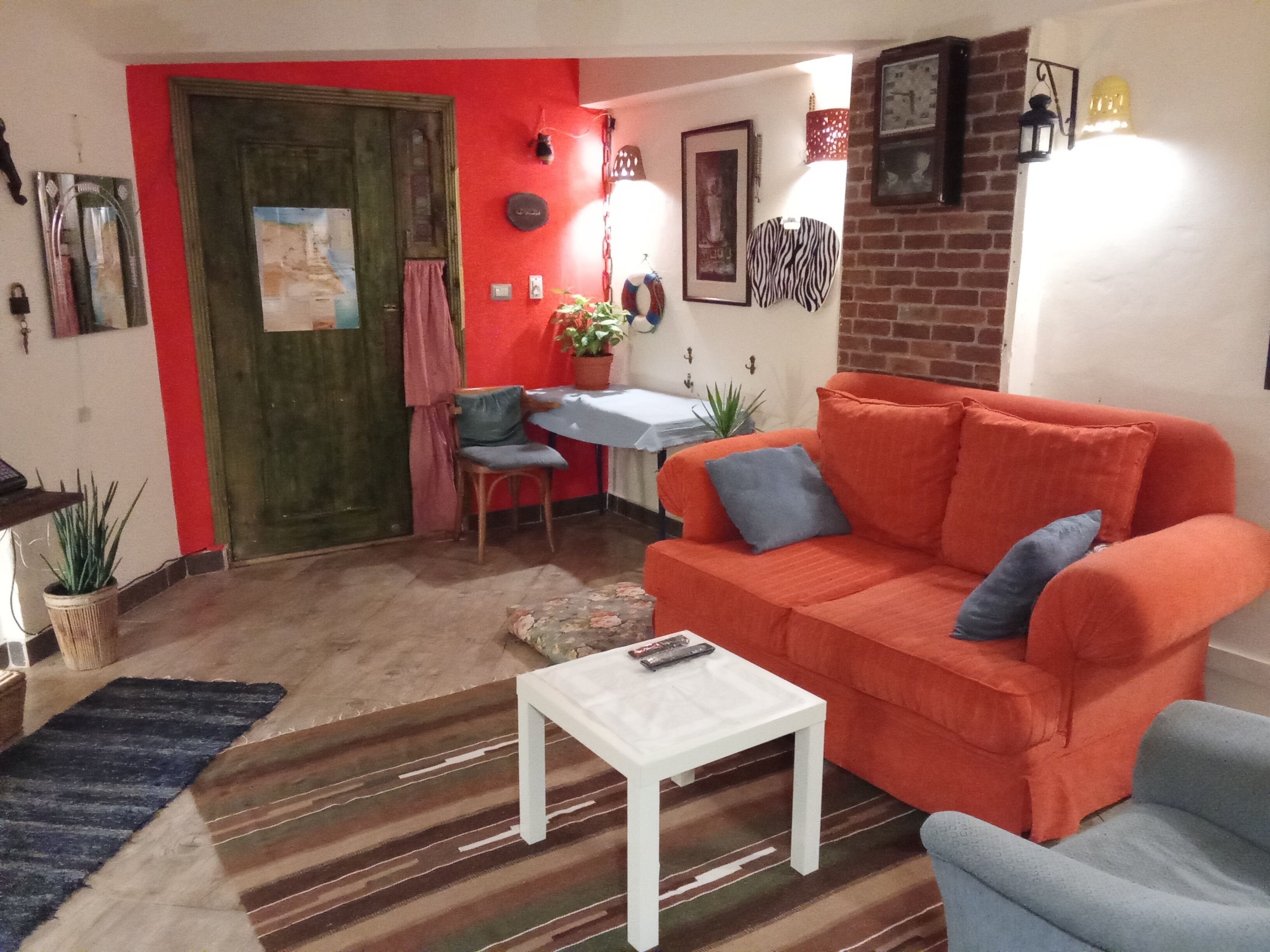 A living room with a red sofa, a white coffee table, and a blue armchair. There is a dining table with chairs and a small potted plant.