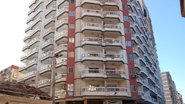A multi-story residential building with balconies, a ground-level shop with closed shutters, and a parked car.