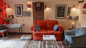 A living room with a red sofa, a blue armchair, a small coffee table, and a patterned rug. There is a brick wall, a clock, and various wall art.