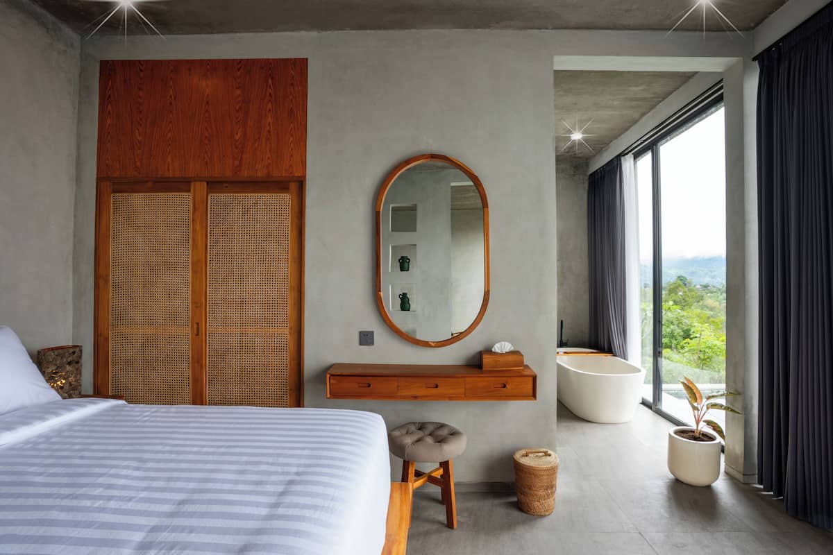 Une chambre moderne avec un grand lit, une armoire en bois, un miroir rond, une table de chevet, un petit tabouret, une plante en pot et une vue sur la verdure grâce à de grandes fenêtres.