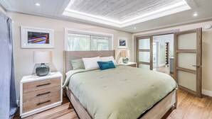 A bedroom with a large bed, a nightstand with a lamp, a dresser, and framed artwork on the wall. - Walk to Harry Harris Beach: Modern Coastal Escape! (Tavernier)