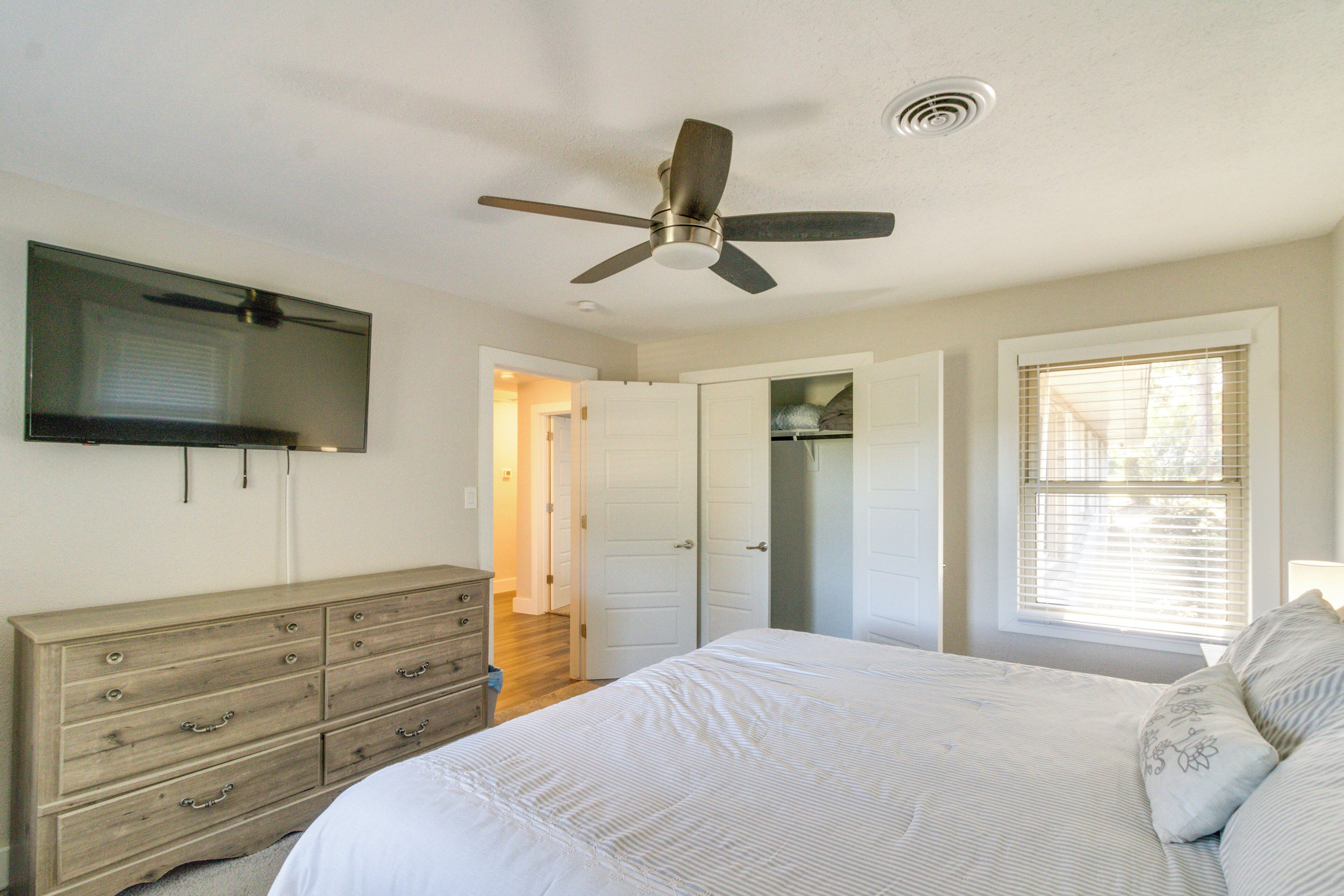 Une chambre à coucher comprenant un lit, une commode, un ventilateur de plafond, une télévision et une fenêtre avec des stores.