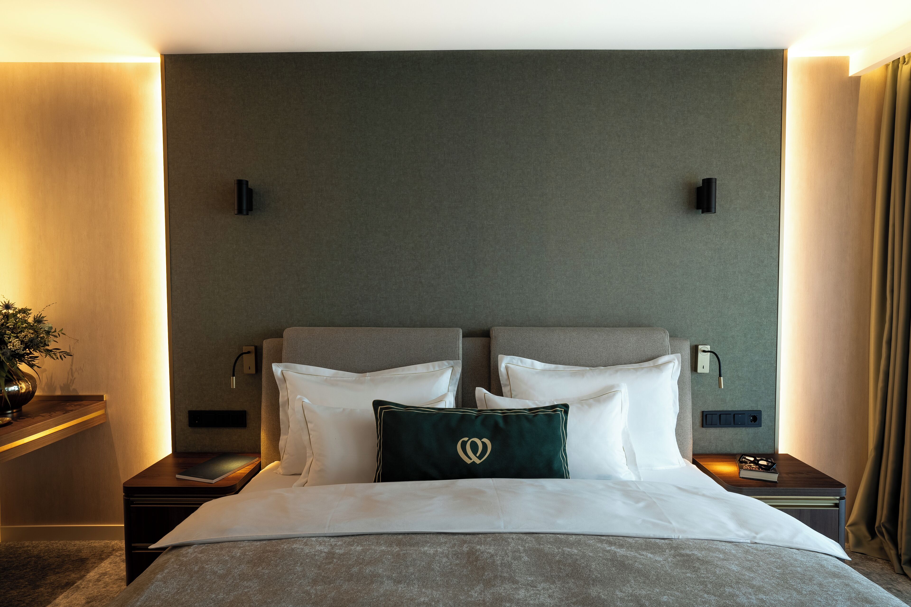 A hotel room with a large bed, two bedside tables, a nightstand, a wall-mounted lamp, and a bedside plant.