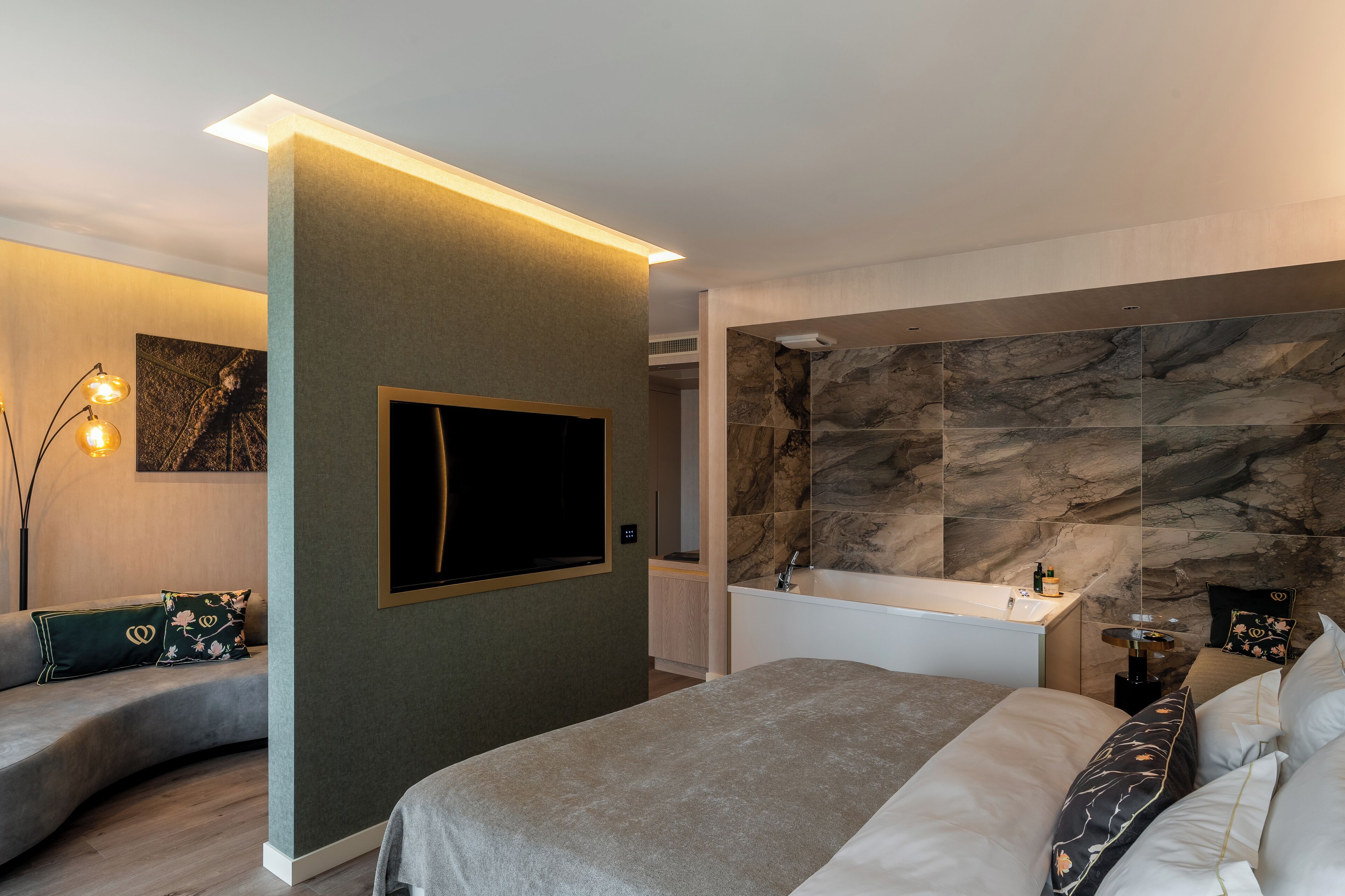 A modern bedroom with a large bed, a built-in TV, a bathroom with a marble wall, and a curved sofa.