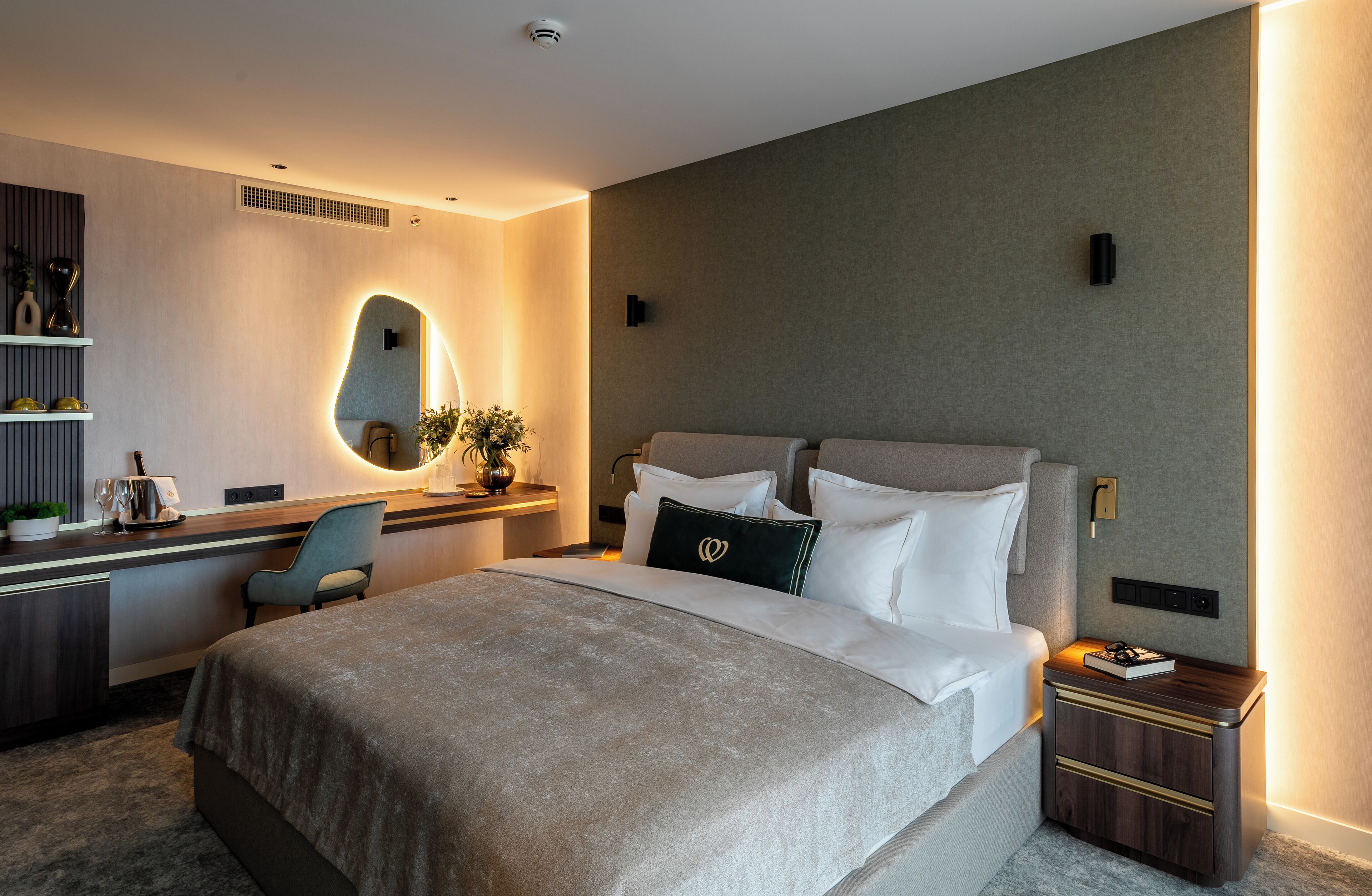 A modern hotel room with a large bed, a desk with a chair, a small table, and a wall-mounted mirror.