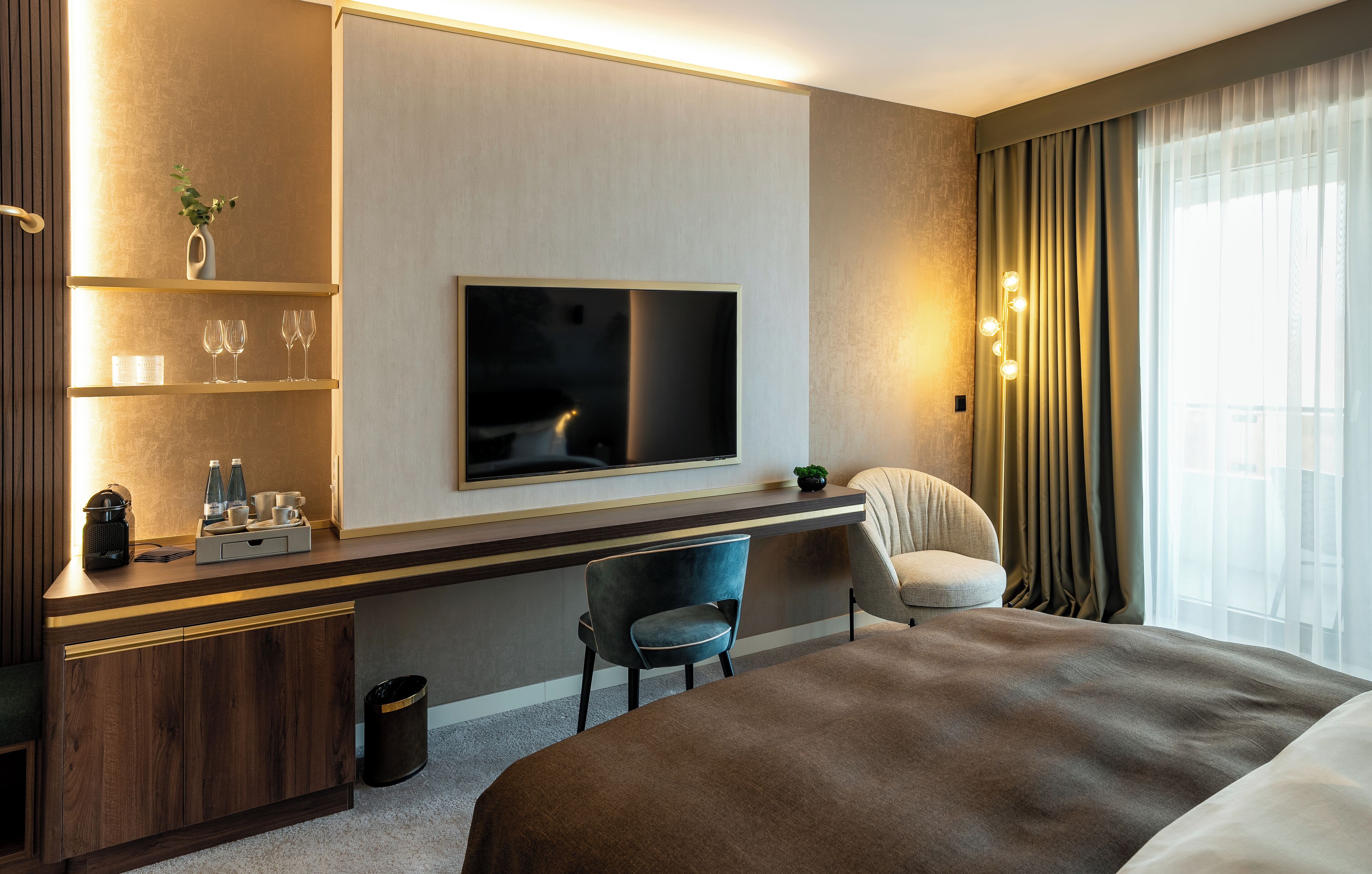 Una habitación de hotel moderna con una cama grande, un escritorio con televisor de pantalla plana, una silla y una pequeña mesa con un florero y vasos.