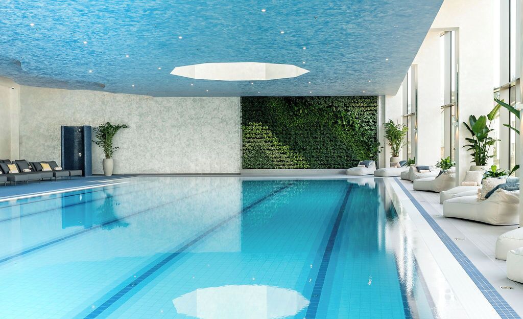 An indoor swimming pool with clear blue water, a ceiling with a geometric light fixture, and a wall with a green vertical garden.