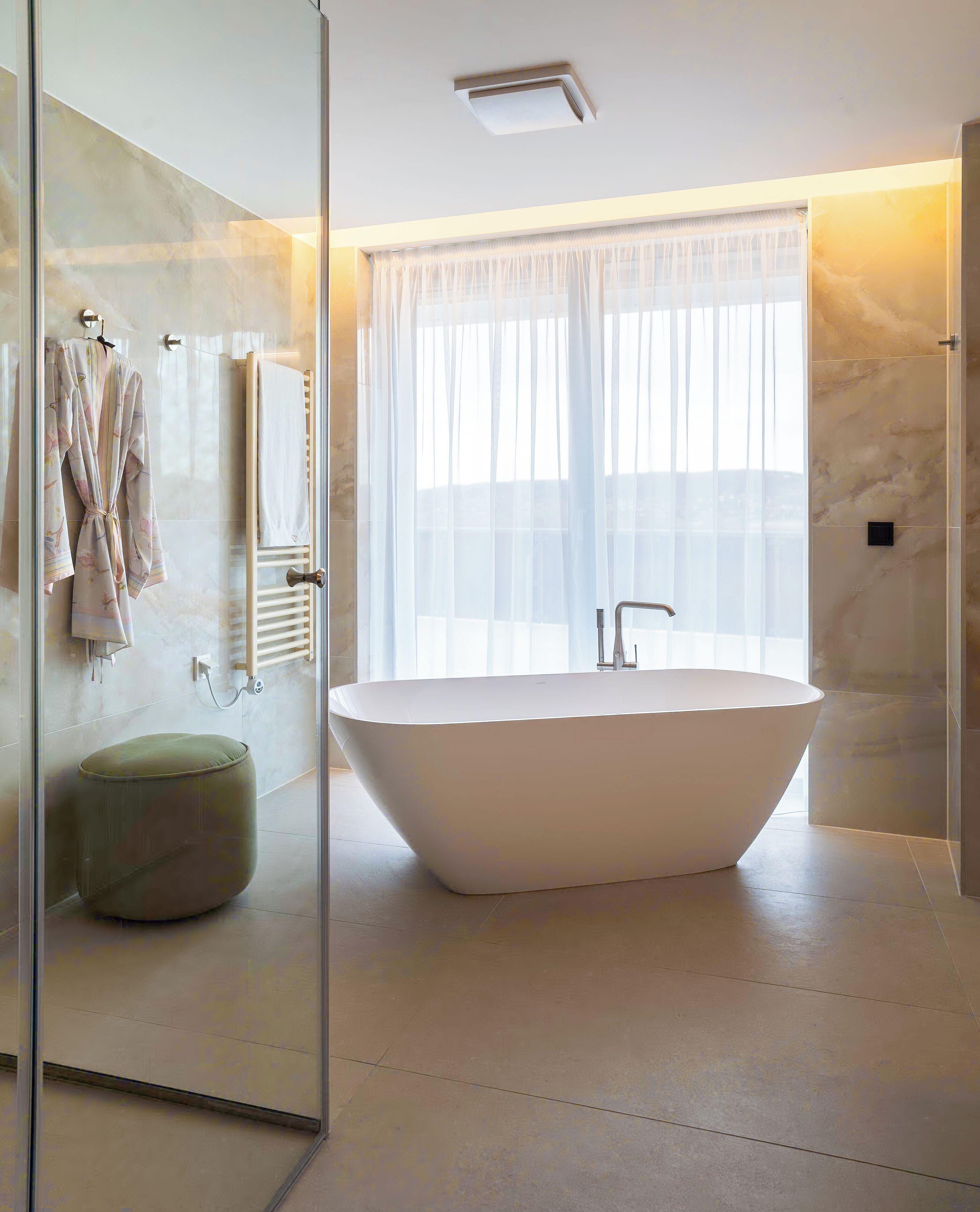 A modern bathroom with a freestanding bathtub, a glass shower enclosure, and a large window with sheer curtains.