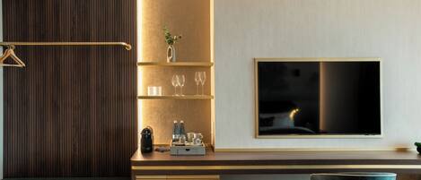 A modern hotel room with a wooden panel wall, a flat-screen TV mounted on the wall, a small seating area with a bench, and a desk with a vase and glasses.