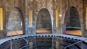 Un área de piscina interior moderna con una piscina circular central, rodeada por pasillos abovedados y una gran lámpara esférica iluminada arriba.