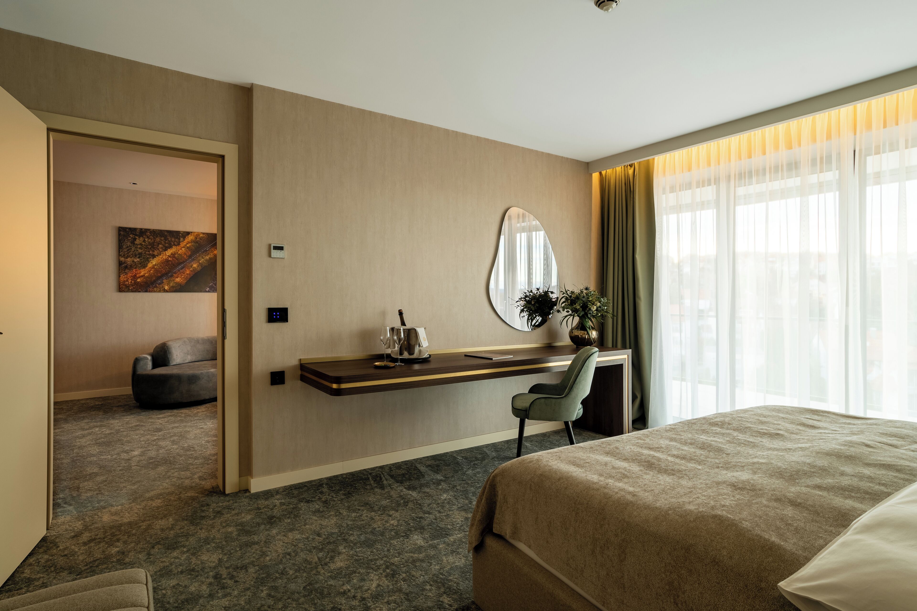 A hotel room with a bed, a desk with a chair, a small sofa, a mirror, and a window with curtains.