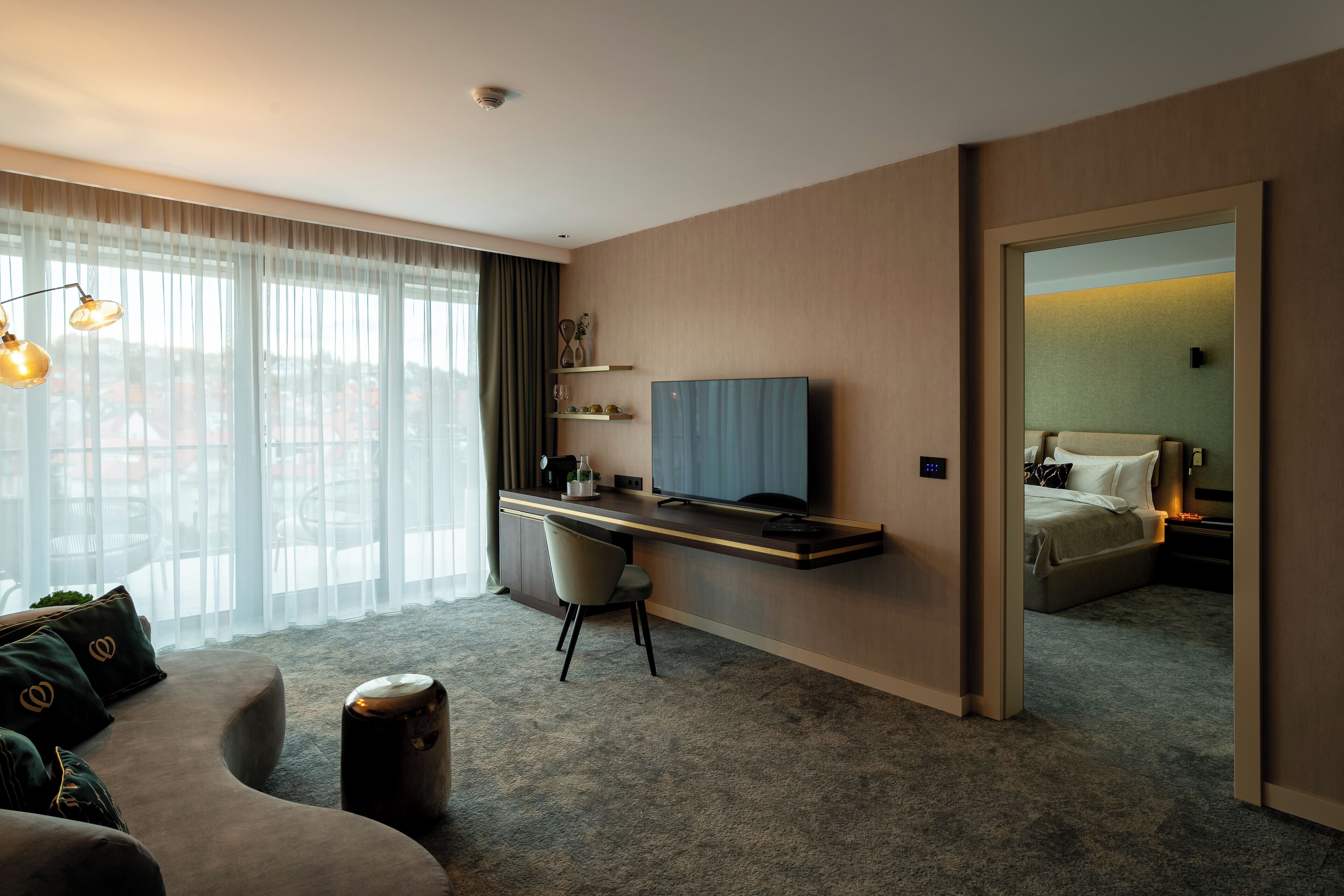 A modern hotel room with a large window, a desk, a comfortable sofa, and a bed.