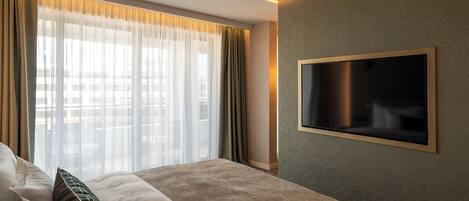 Habitación de hotel con una cama grande, un televisor de pantalla plana en la pared y vistas de la ciudad a través de cortinas translúcidas.