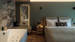 Un dormitorio moderno con una cama grande, una zona de estar separada con un banco acolchado, una mesita de noche pequeña con lámpara y una bañera grande.