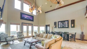 A spacious living room with a large sofa, a coffee table, and a dining area with chairs and a table. There are large windows with blinds, a ceiling fan, and artwork on the walls. - Beachfront Group Getaway on Bolivar Peninsula (Port Bolivar)