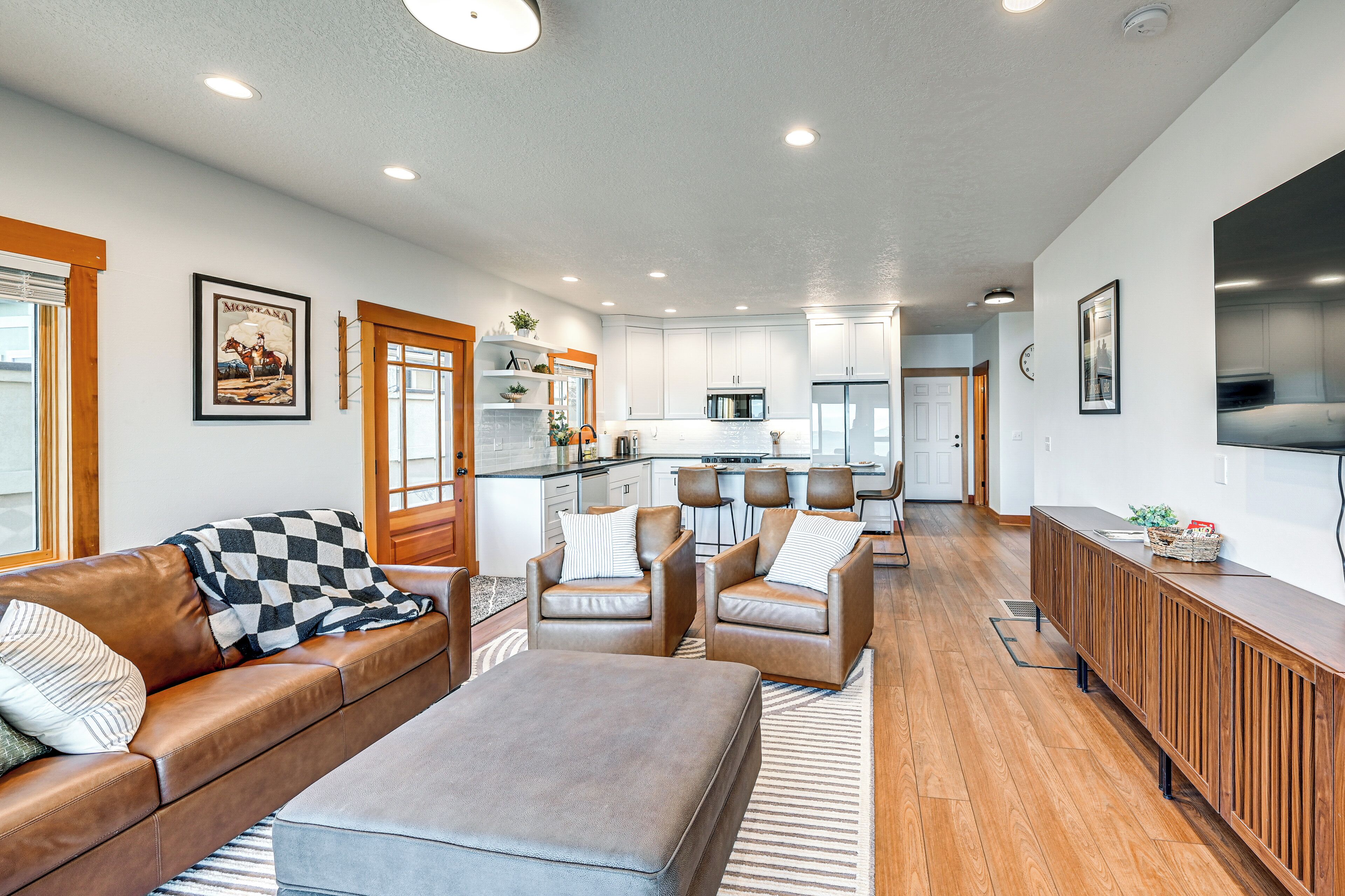 A spacious living area with a brown leather sofa, two armchairs, and a wooden coffee table. A kitchen with white cabinets and a central island is visible in the background.