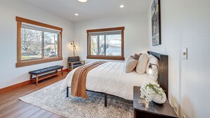 Une chambre à coucher avec un grand lit, deux fenêtres, un banc et une table de chevet ornée d’un vase de fleurs.