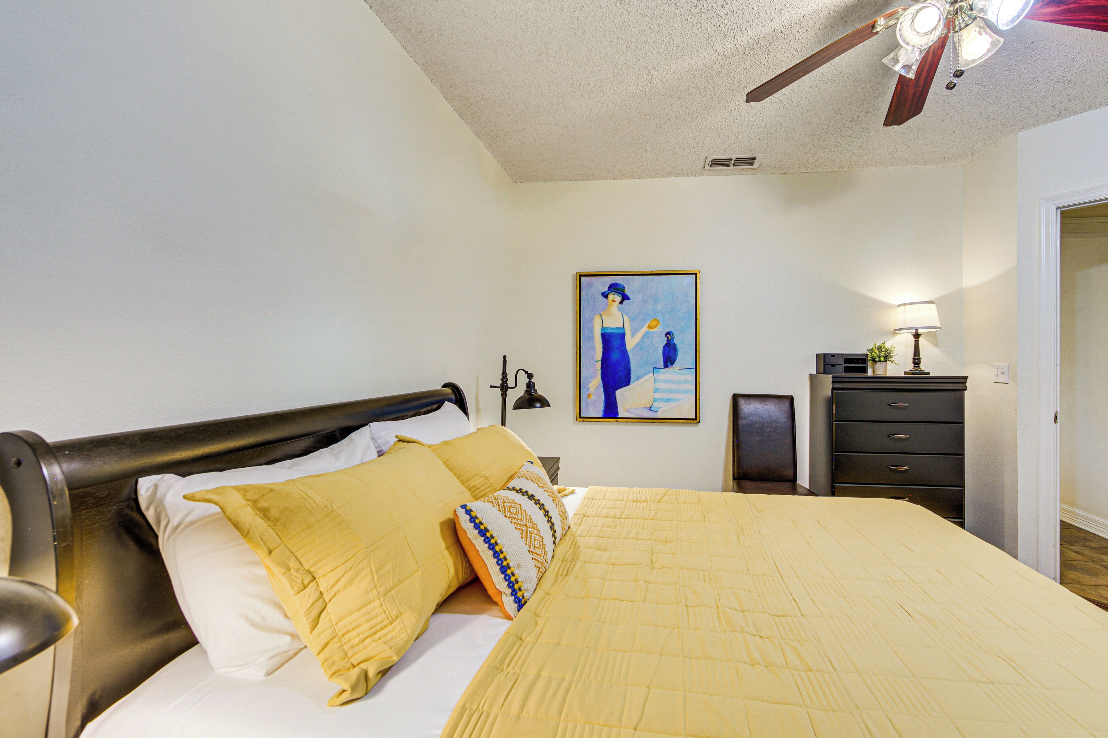A bedroom with a bed, a nightstand, a dresser, a ceiling fan, and a painting on the wall.