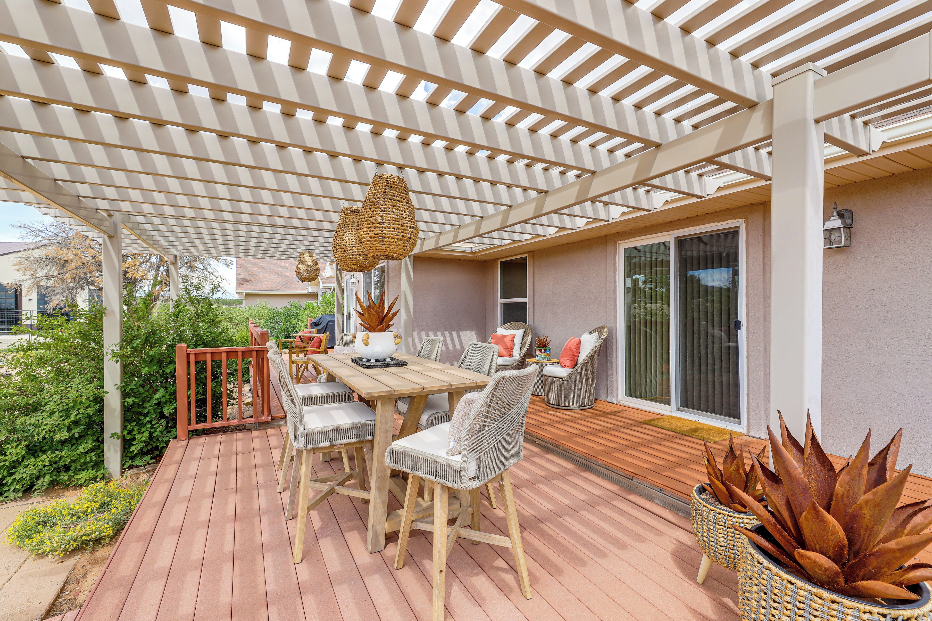 A covered outdoor patio with a wooden table and chairs, a woven hanging light fixture, and a potted plant.