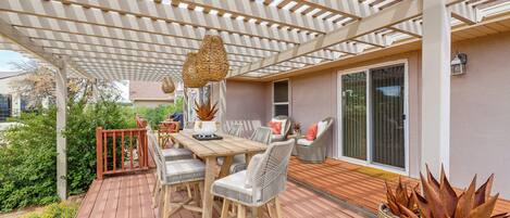 A covered outdoor patio with a wooden table and chairs, a woven hanging light fixture, and a potted plant.