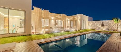A modern outdoor pool area with a wooden deck, a stone wall, and a well-lit interior.