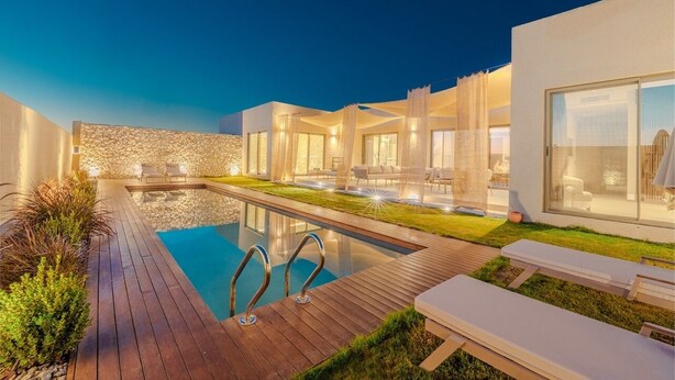 A modern outdoor pool area with a wooden deck, lounge chairs, and a stone wall. - Silence Villas (Fethiye)
