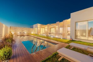 A modern outdoor pool area with a wooden deck, lounge chairs, and a stone wall. - Silence Villas (Fethiye)