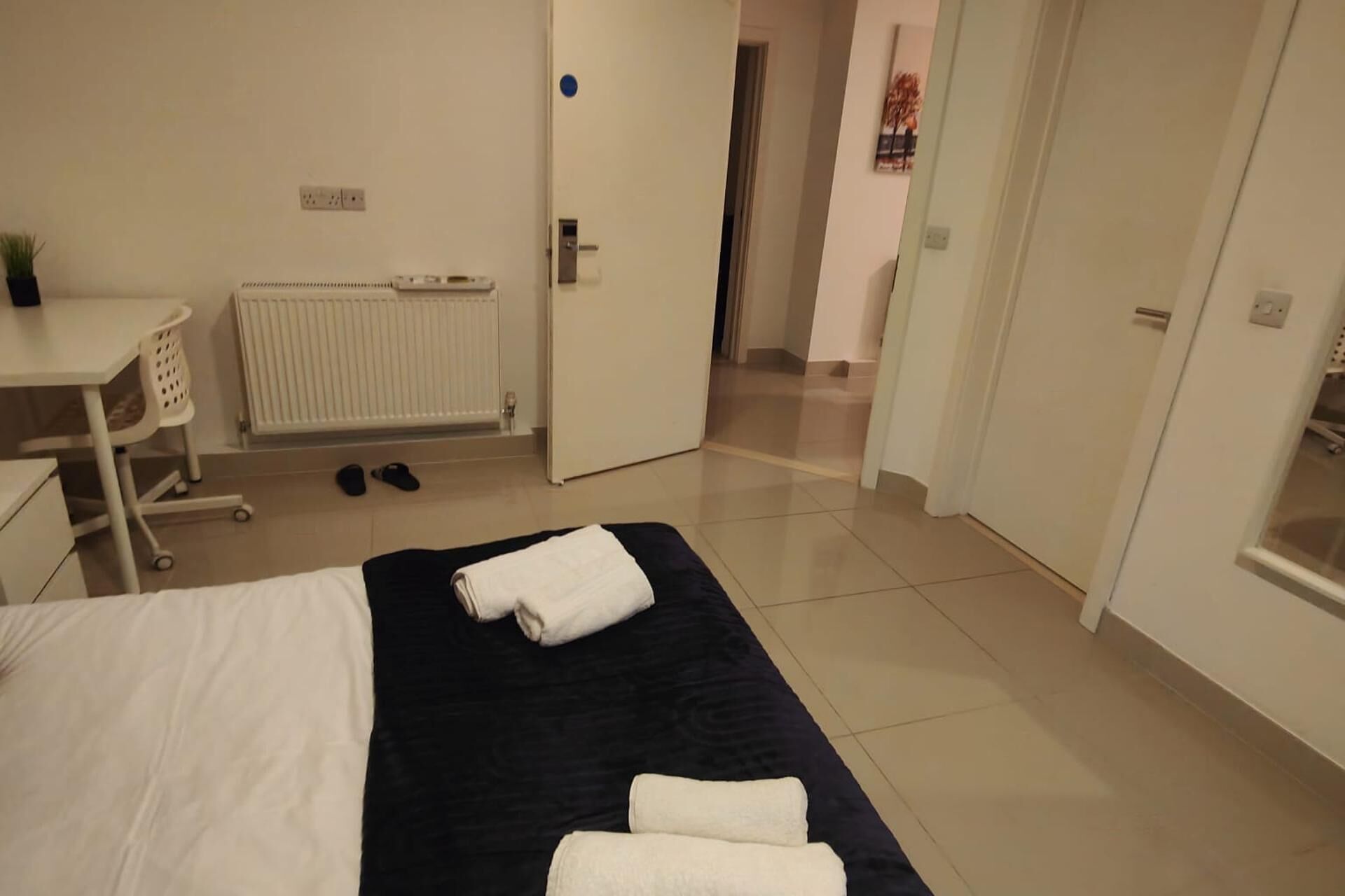 Een hotelkamer met een bed, een bureau, een radiator en een deur naar een andere kamer.