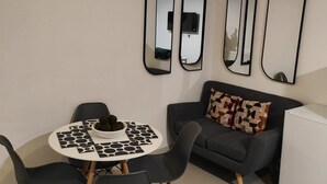 Uma sala de estar moderna com uma mesa de jantar redonda, cadeiras pretas, um sofá cinza com almofadas estampadas e três espelhos fixados na parede.