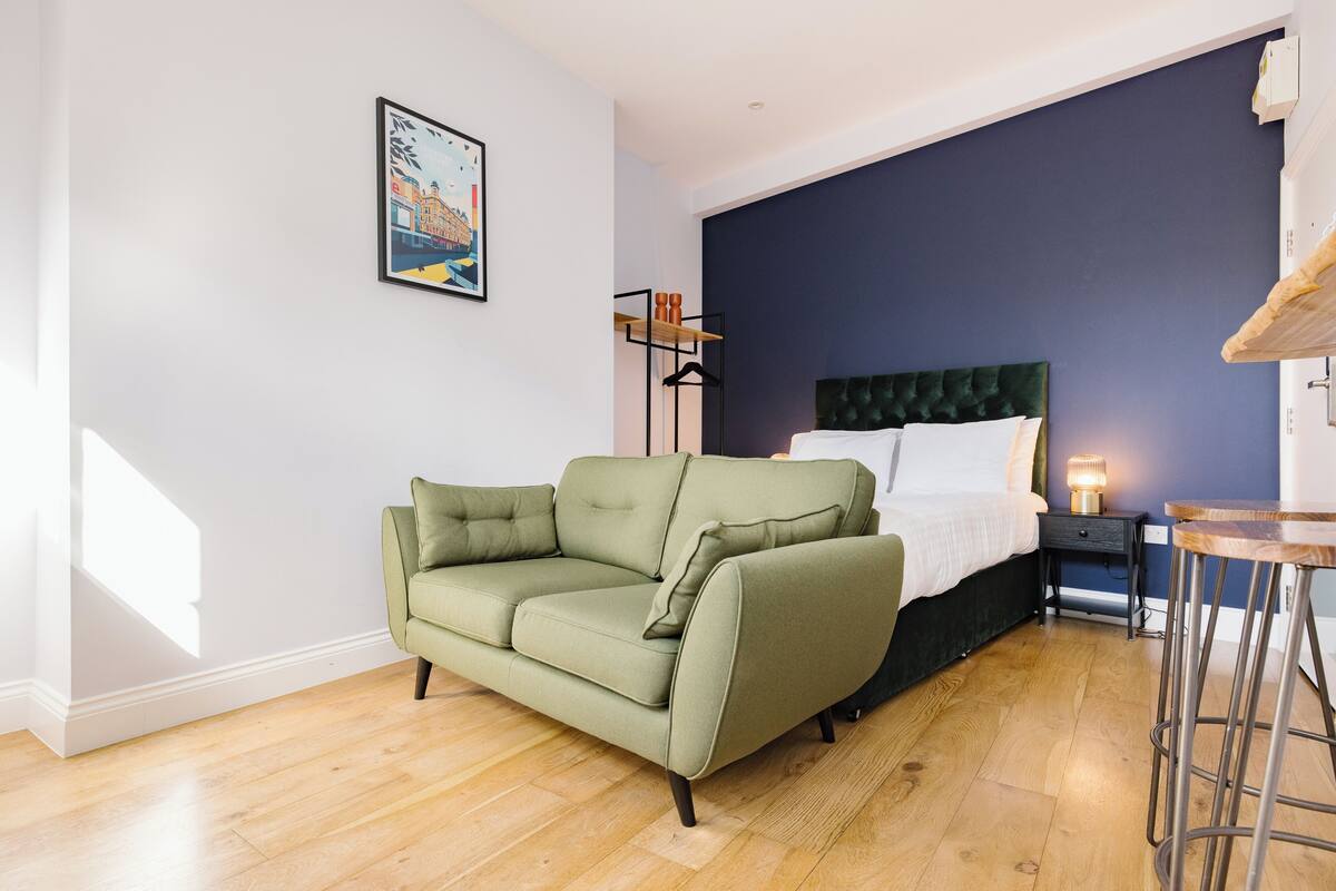 A modern bedroom with a green sofa, a bed with a dark headboard, a nightstand with a lamp, and a wooden stool.
