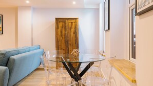 Een moderne woonkamer met een eettafel met glazen blad, omringd door transparante stoelen, een blauwe bank en een houten deur op de achtergrond.
