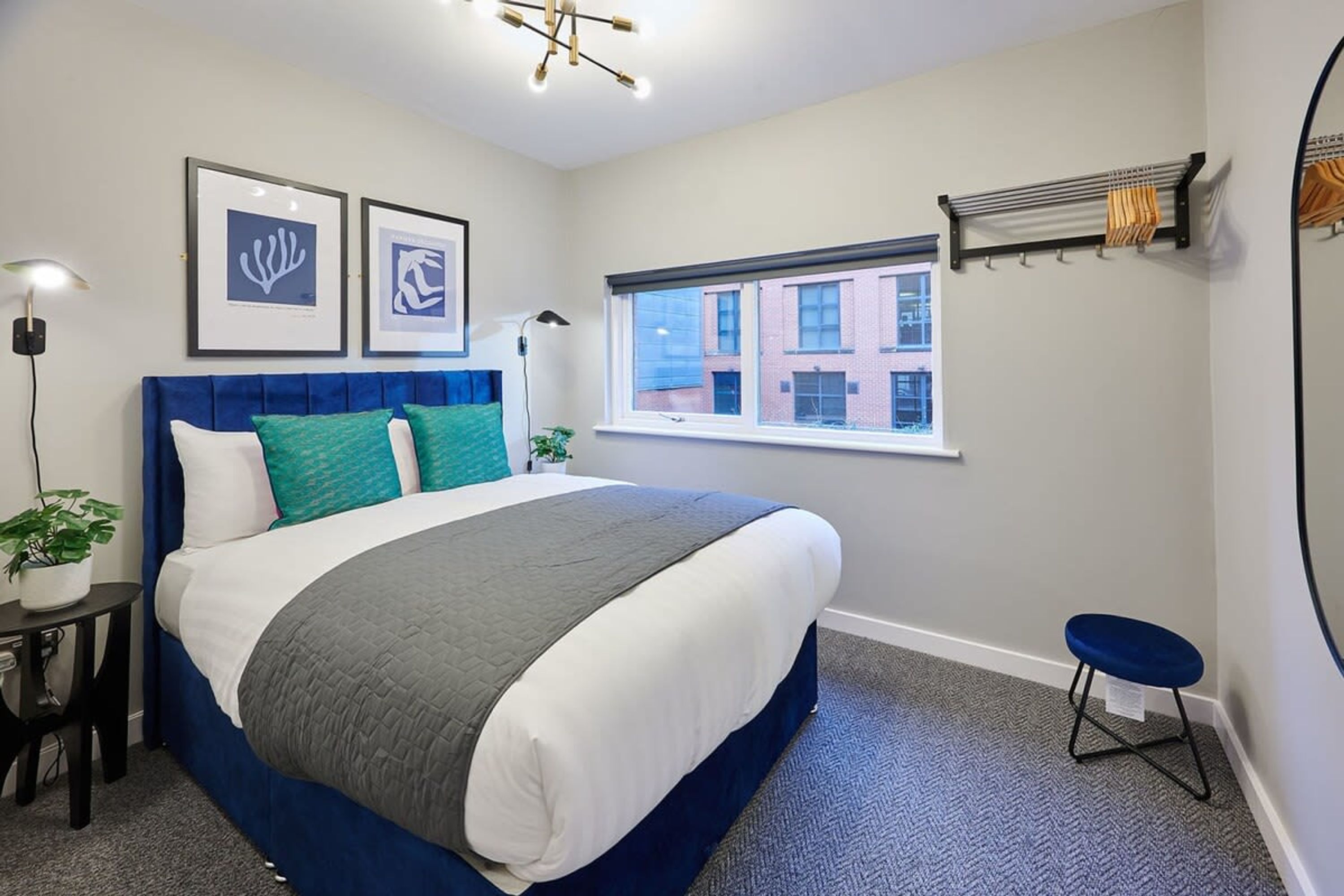 Een slaapkamer met een groot bed, een raam met uitzicht op gebouwen, een nachtkastje met een lamp en een klein krukje.