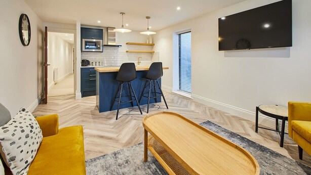 Een moderne woonkamer met een houten salontafel, een gele bank en een keukenhoek met een barkruk.