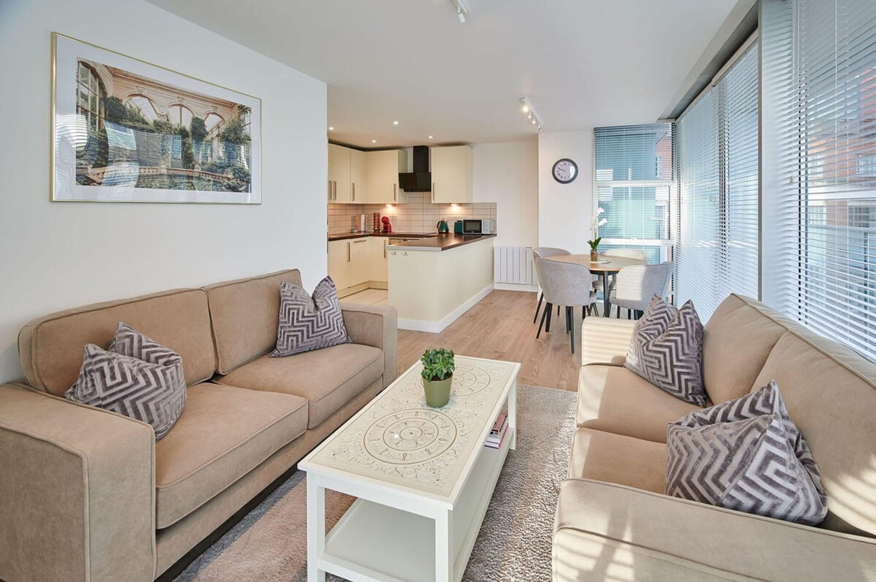 A modern living room with a beige sofa set, a coffee table with a decorative top, and a dining area with a small table and chairs. A kitchen is visible in the background with white cabinets and a black range hood.