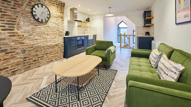A modern living room with a brick wall, a round clock, a green sofa, a wooden coffee table, and a kitchen in the background. - Host Stay Sea Salt Apartment (Whitby)