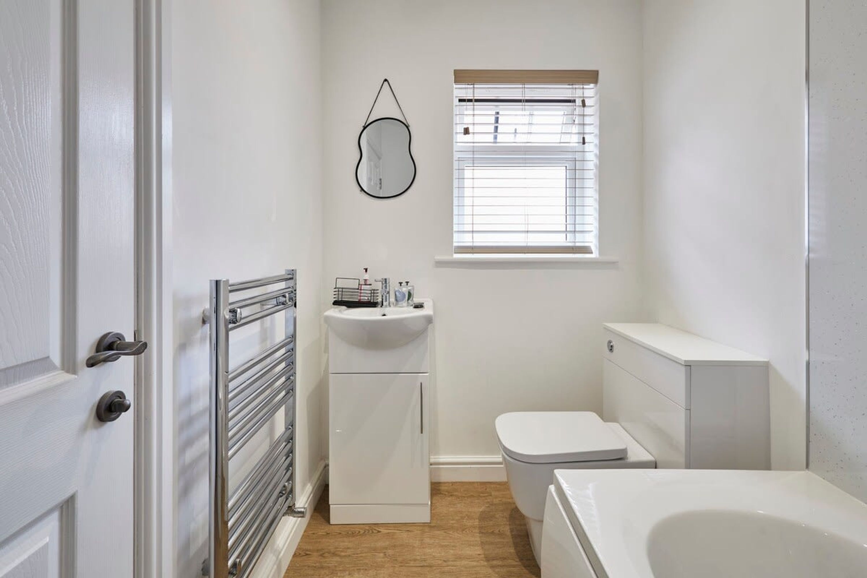 Une salle de bain compacte avec un lavabo, des toilettes et une baignoire, tous de couleur blanche. Une fenêtre équipée de stores laisse entrer la lumière naturelle.