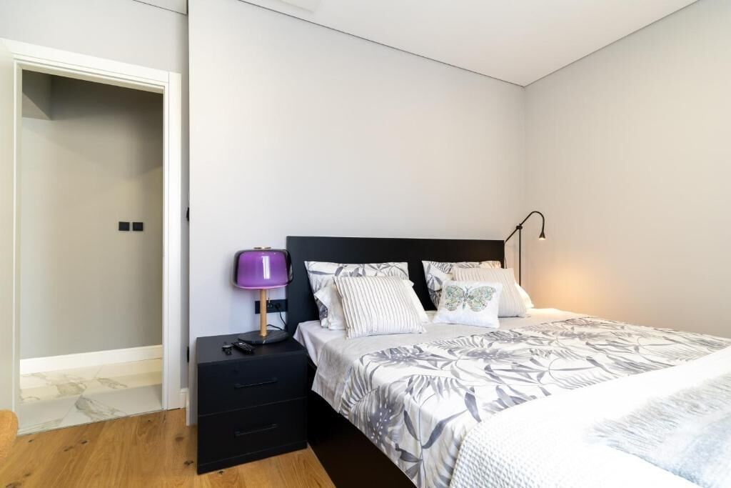 A bedroom with a bed, a nightstand with a lamp, a drawer unit, and a door leading to another room.