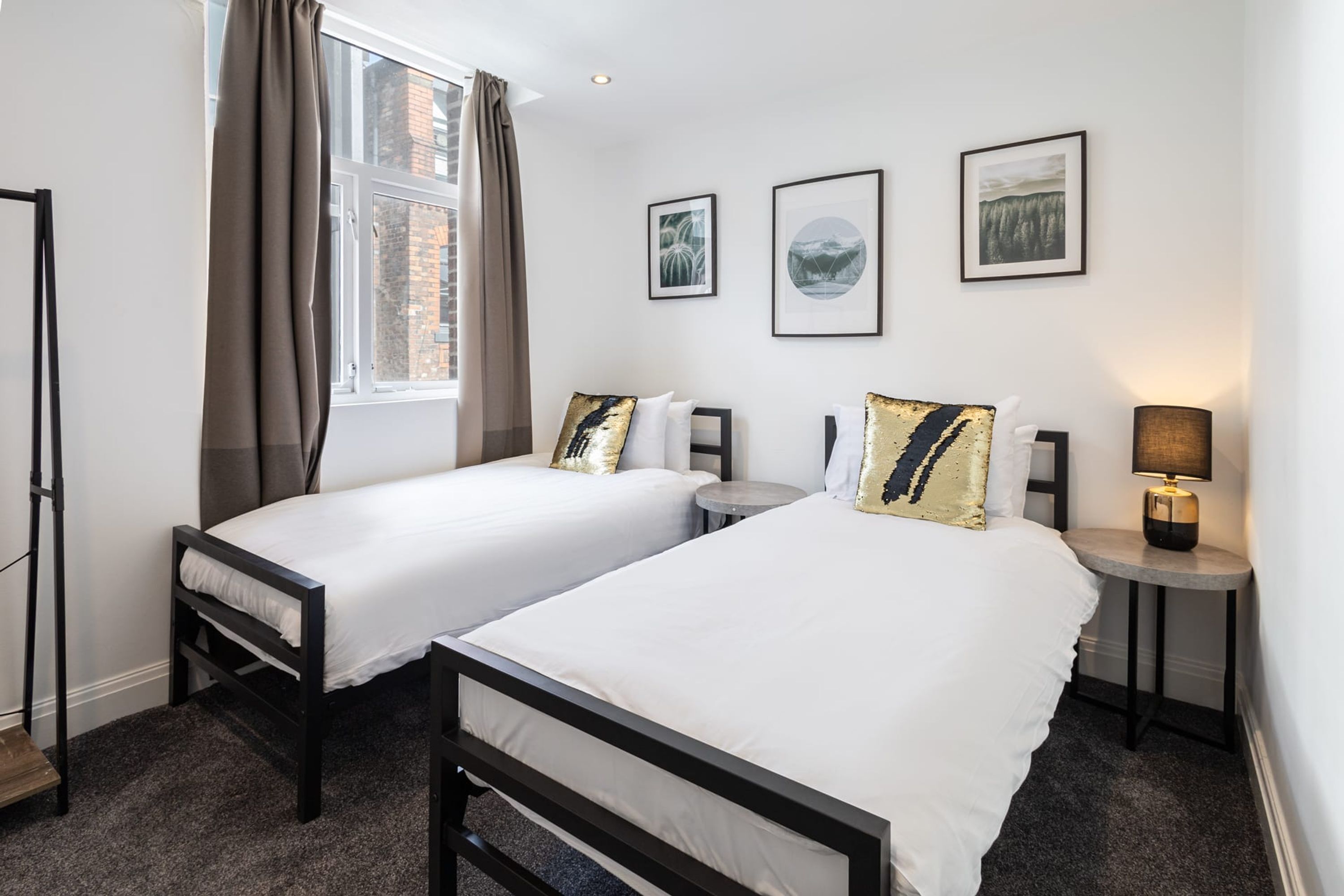 Habitación de hotel con dos camas, una ventana con cortinas, una mesita de noche con lámpara y cuadros enmarcados en la pared.