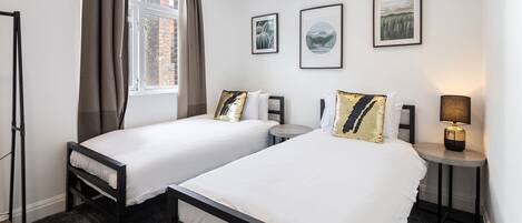 Habitación de hotel con dos camas, una ventana con cortinas, una mesita de noche con lámpara y cuadros enmarcados en la pared.