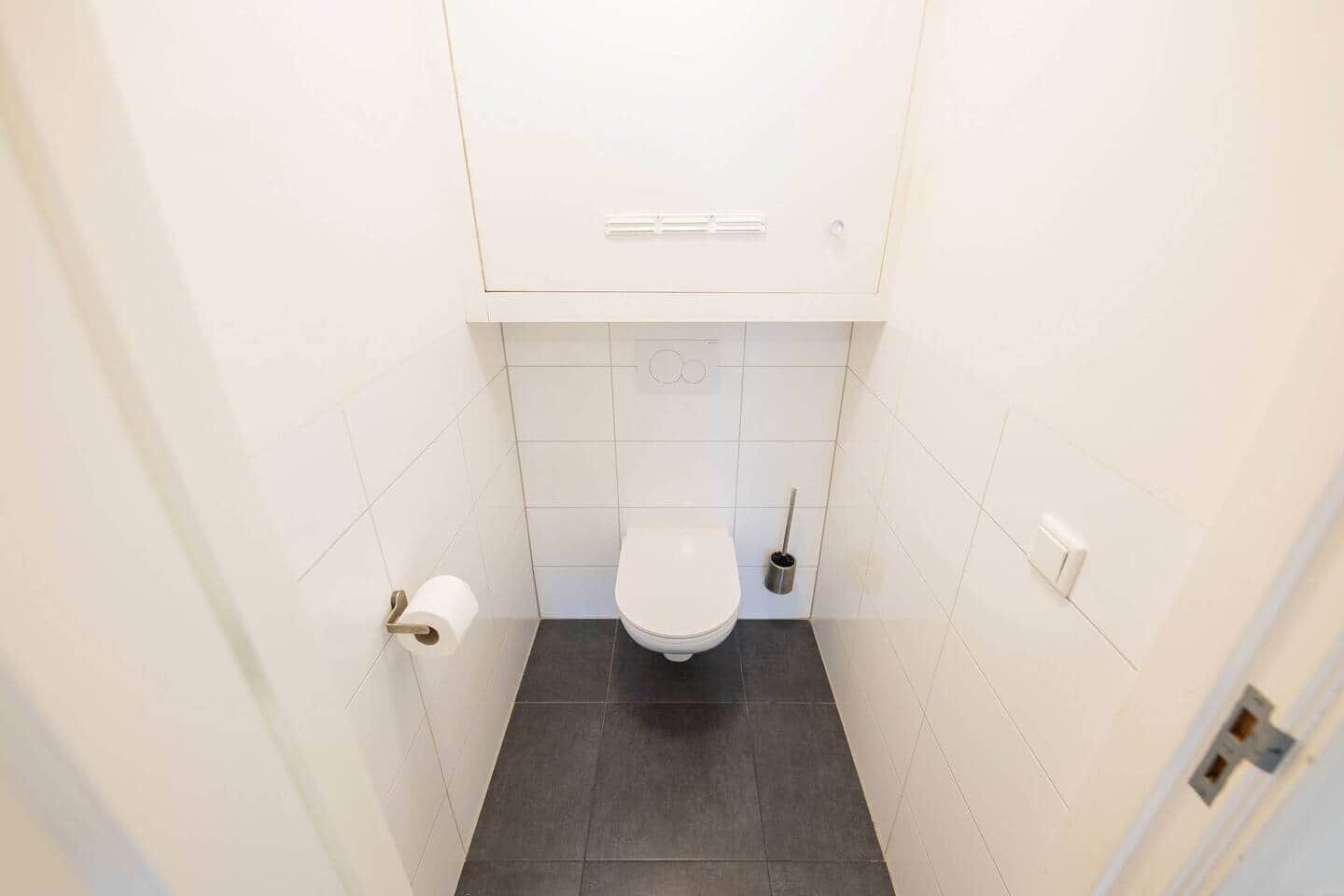 A small, narrow bathroom with a toilet, a toilet paper holder, and a broom.