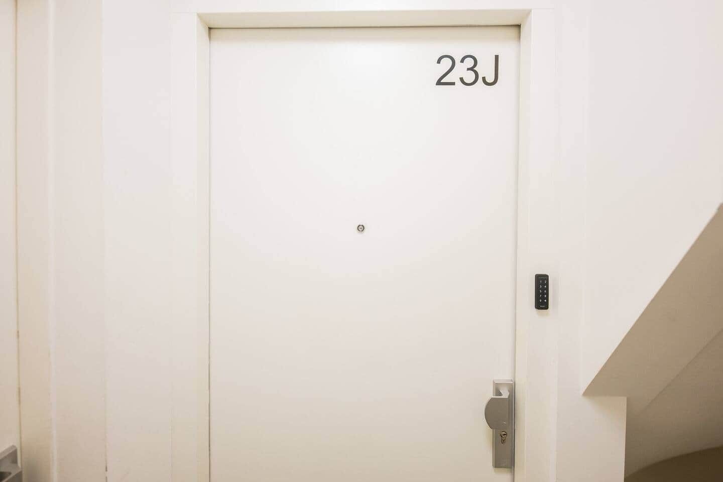 En vit dörr med numret 23J på, ett lås med knappsats och ett litet handtag.