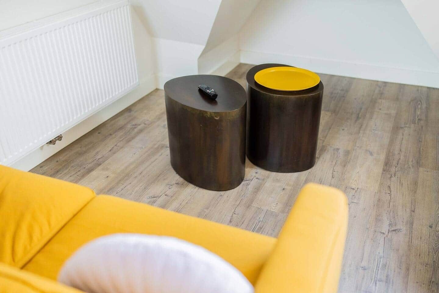 Ein Zimmer mit Holzboden, einem weißen Heizkörper und zwei dunklen, zylindrischen Objekten mit gelben Spitzen.