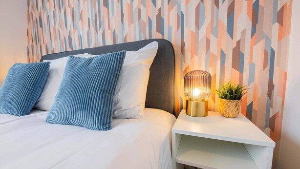 Una cama bien hecha con almohadones azules, una mesita de noche con una lámpara y una planta, y una pared con un diseño geométrico.