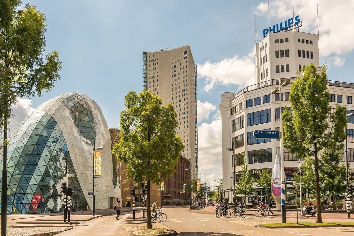 Una strada cittadina moderna con un caratteristico edificio in vetro, un edificio per uffici Philips, e un marciapiede alberato.