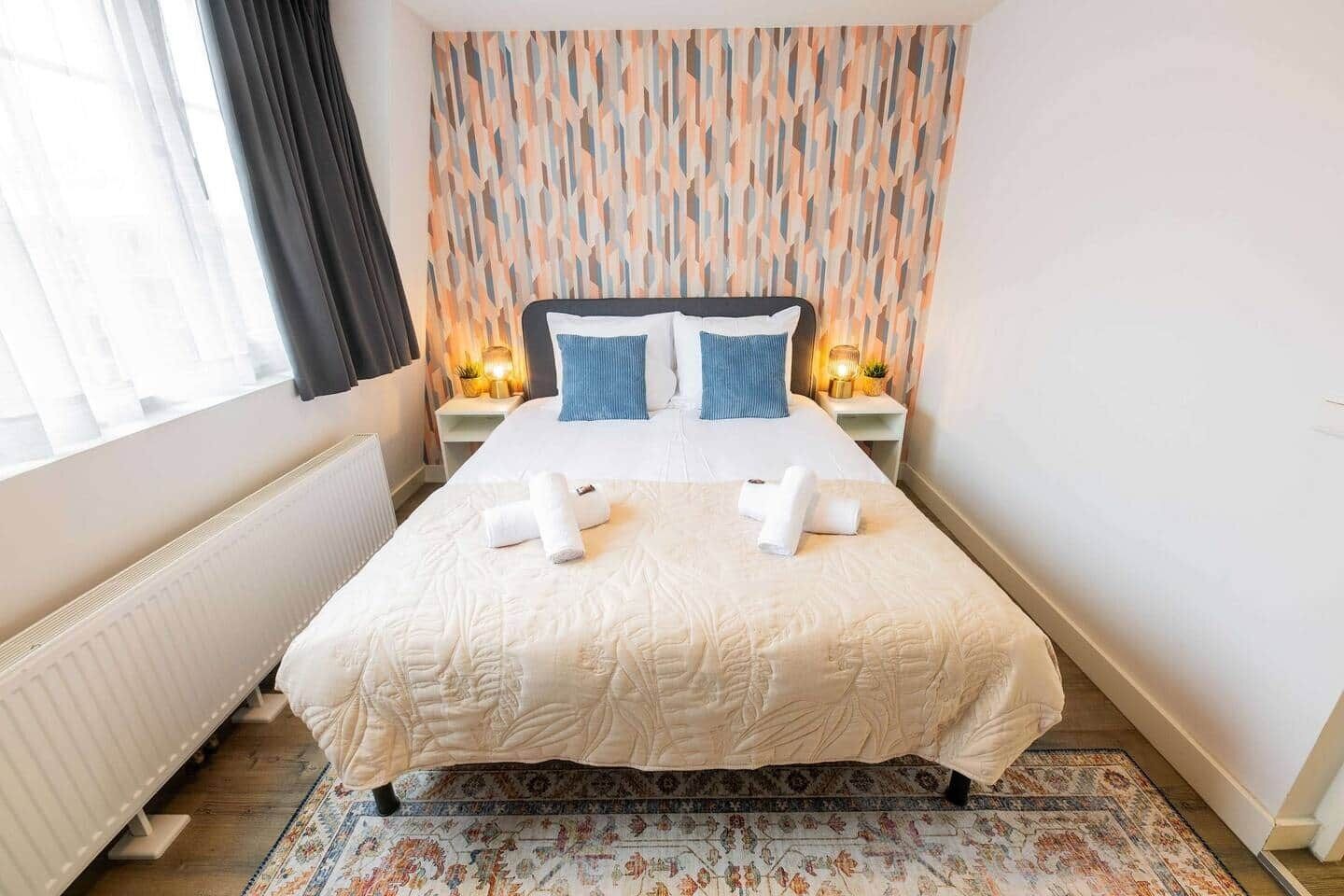 Un lit bien fait, avec une couette blanche et deux oreillers, un tapis à motifs et un mur orné d’un motif géométrique.